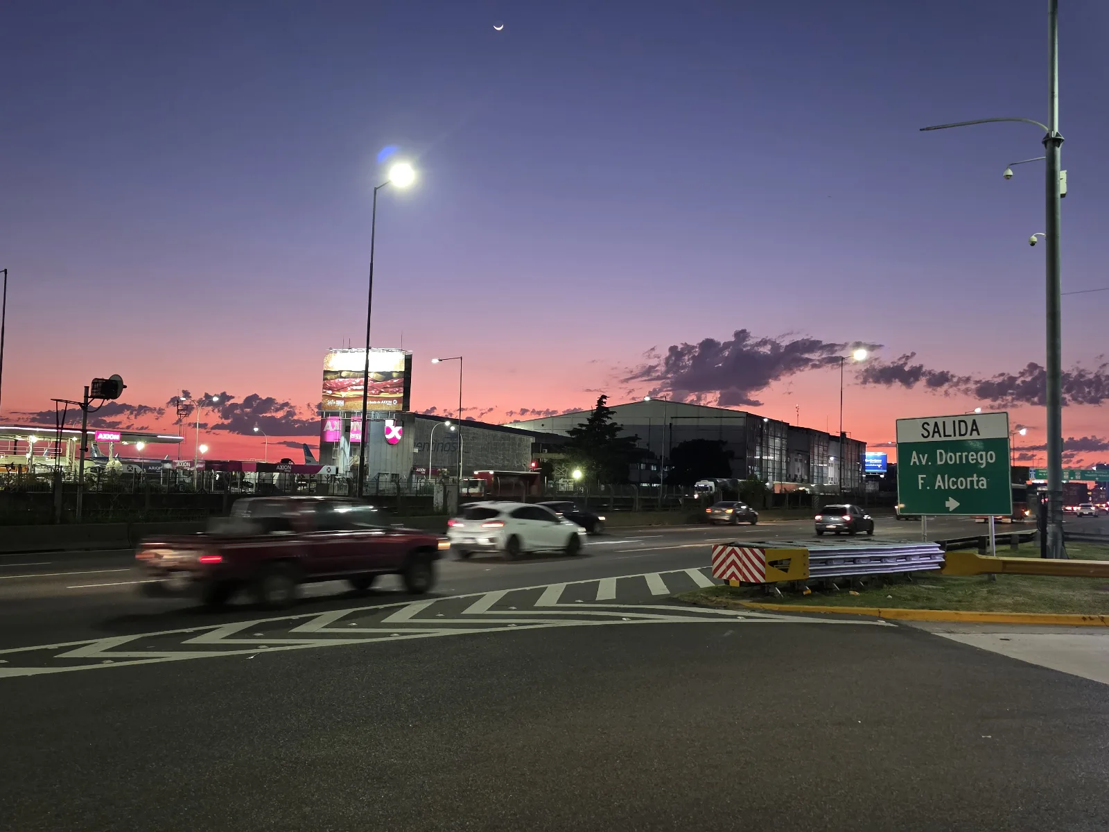 View of the fuel sector and perimeter area of Aeroparque Jorge Newbery (AEP/SABE) at dawn, Buenos Aires, Argentina. In the foreground is Avenida Costanera Rafael Obligado with moving traffic (a red pi